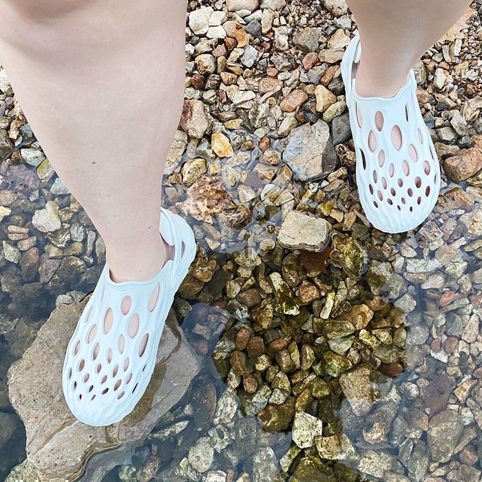 A pair of white, perforated shoes rests on wet stones in shallow water, with a partial view of a bare leg visible.