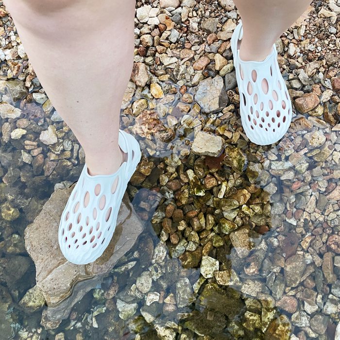 A person stands in shallow water, wearing white, hole-patterned shoes, with exposed legs on a rocky riverbed.