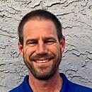 A man with a short beard and short hair smiles, wearing a blue shirt, against a textured light-colored wall.