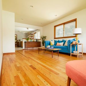 A colorful sofa with pillows sits on polished wood flooring. A coffee table is nearby, and a lamp stands beside the couch in a bright room.