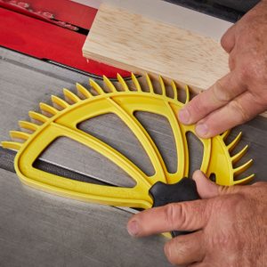 A yellow push stick is positioned against a piece of wood, assisting in guiding it through a table saw's blade in a workshop setting.