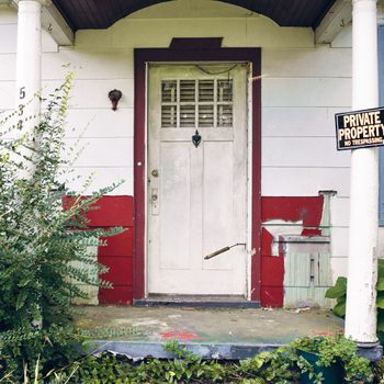 Old, Abandoned House with Private Property Sign Posted on the Front Porch. The Home looks run down and abandoned with overgrown weeds and trees, chipping paint on the siding and around the front door.