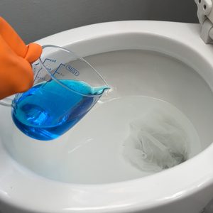 A person wearing orange gloves is pouring a blue liquid from a measuring cup into a toilet. The toilet seat is lifted.