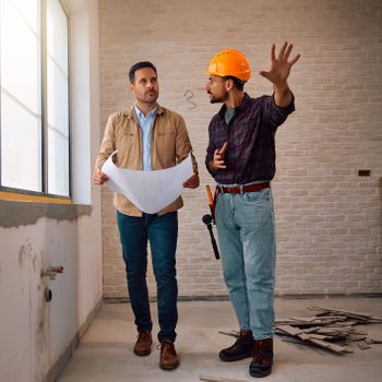 Home owner and contractor talking while analyzing blueprints at construction site.